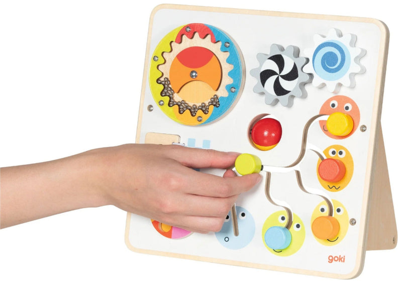 A Goki wooden sensory and motor activity board with colorful gears, buttons, and animal faces, shown with a child's hand interacting with the toy on a white background.