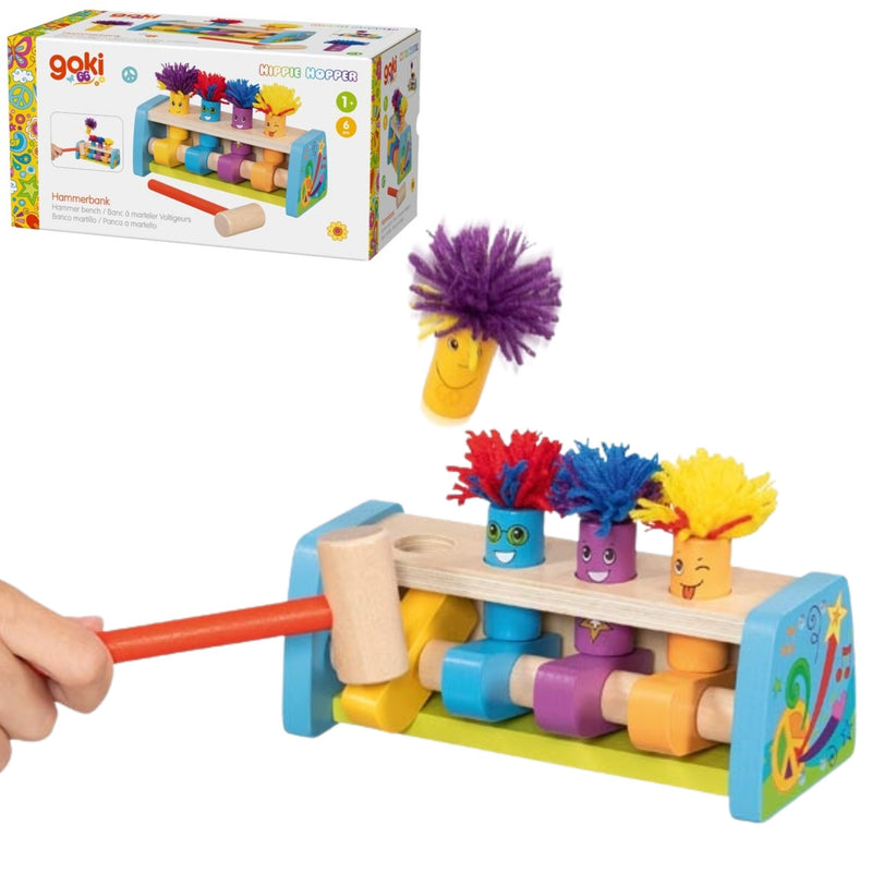 A children's wooden toy set shown on a white background, including a colorful peg-bobbing frame with three smiling character tops, a wooden mallet being swung by a visible hand, a loose purple pom-pom, and the retail box in the upper left. The brand name Gaki is visible on the packaging.