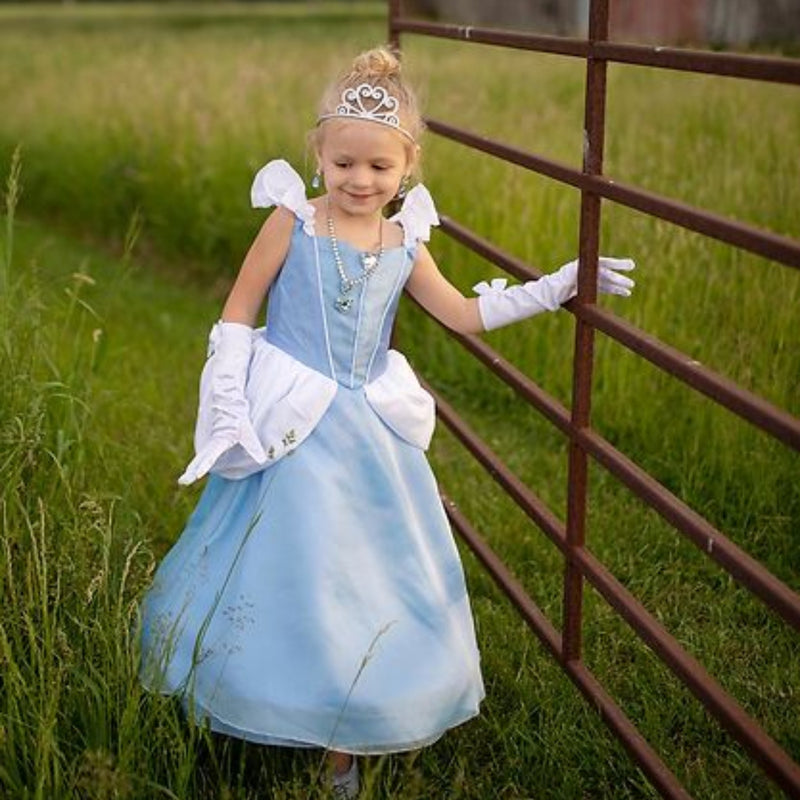 A young girl wearing a light blue princess costume dress with white puffed sleeves and gloves, a tiara, and a necklace, standing in a grassy field next to a metal gate.