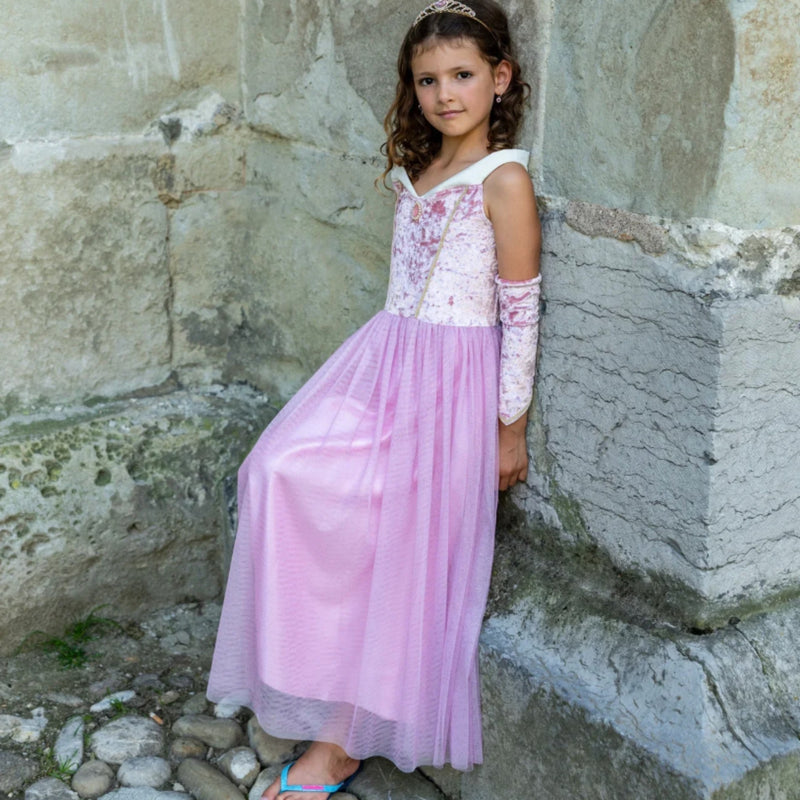 A young girl with curly brown hair stands against a rustic stone wall wearing a light pink and white princess costume with a long pink sleeve and blue flip-flops.