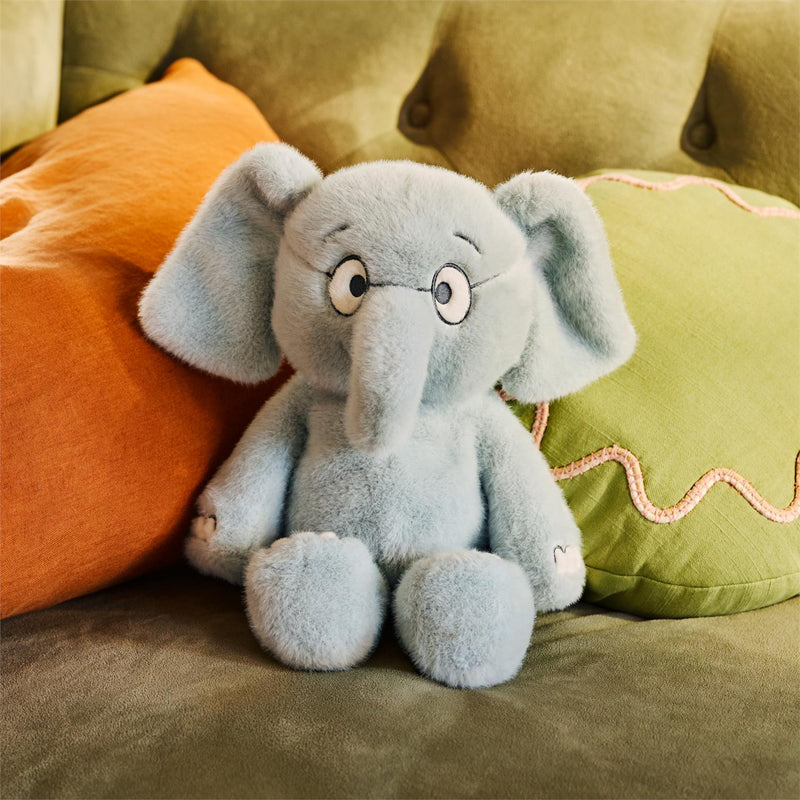 Mo Willems' Gerald elephant plush seated on a couch with colorful pillows