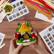 Hands holding completed chenille mushroom house craft with colorful materials and instruction sheets