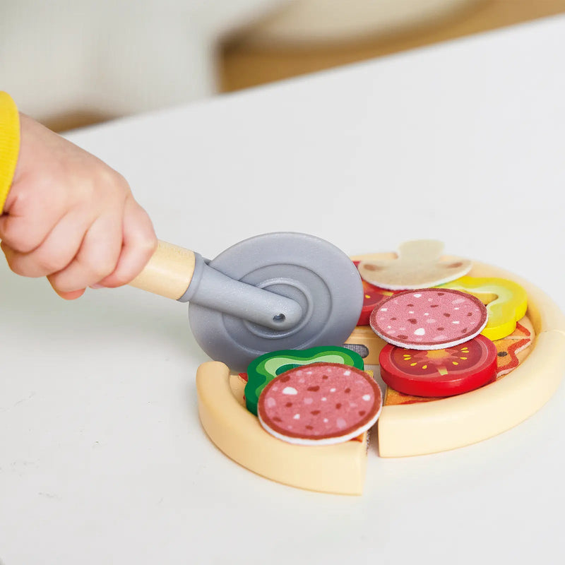Child's hand using toy pizza cutter on wooden play pizza with colorful toppings