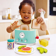 Toddler joyfully playing with Hape mint green pop-up toaster toy set with accessories