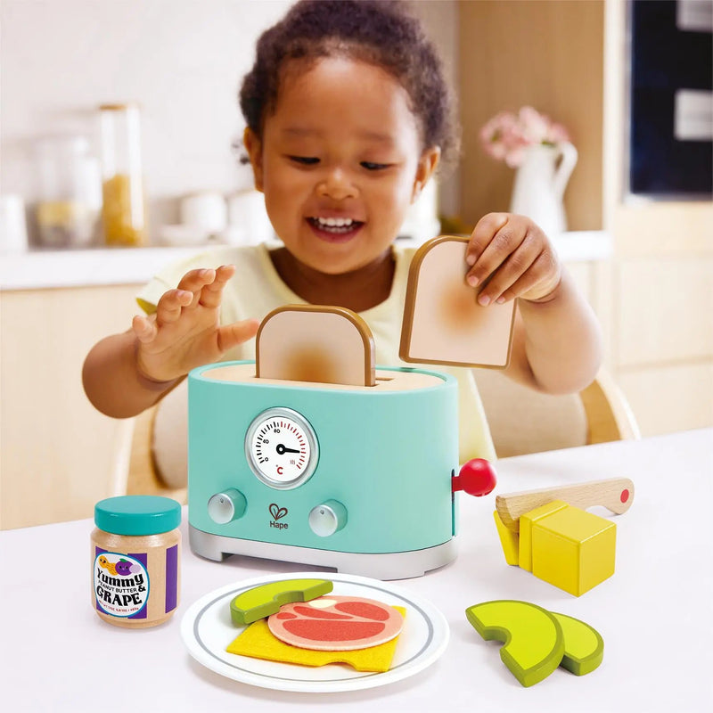 Toddler joyfully playing with Hape mint green pop-up toaster toy set with accessories