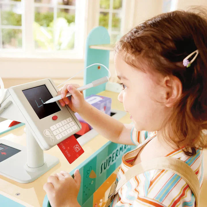 Young girl playing cashier signing toy payment terminal at Hape wooden supermarket set