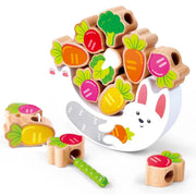 The Bunny Balance game with colorful vegetable and animal pieces on a white background. The set includes carved wooden blocks with painted radishes, carrots, broccoli, and a caterpillar, plus a white bunny-shaped tray or holder with pink inner ears and a gray slide.