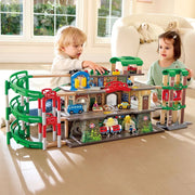 A children's wooden multi-level toy train station and track set with colorful tracks, cars, and small figures, shown in a bright living room with two children playing around it.