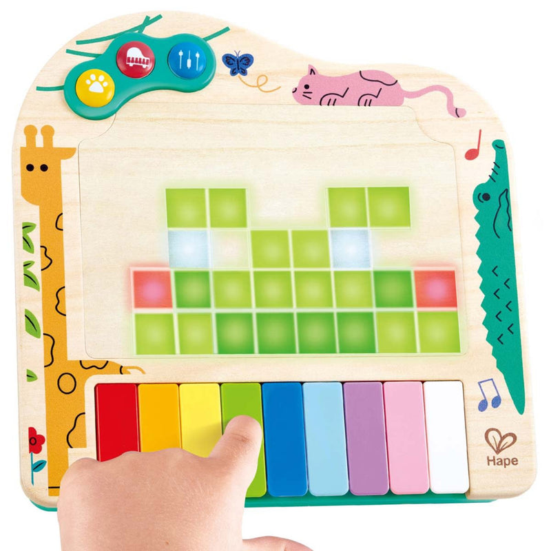 Hape's Pixel Piano wooden musical toy with a colorful keyboard, touch-sensitive green and red blocks, animal illustrations, and a child's hand pressing the keys. The brand name 'Hape' is visible in the bottom right corner.