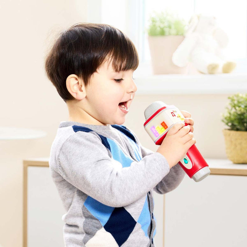 A young child wearing a gray and blue argyle sweater holds a red and white toy microphone, singing with an open mouth in a brightly lit indoor room.