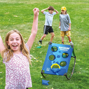 A girl in a pink floral shirt celebrates in the foreground with her arm raised, while two boys play on a blue Rockin' Pockets portable target game on a green grass lawn. The boy on the right throws a bean into the pockets.
