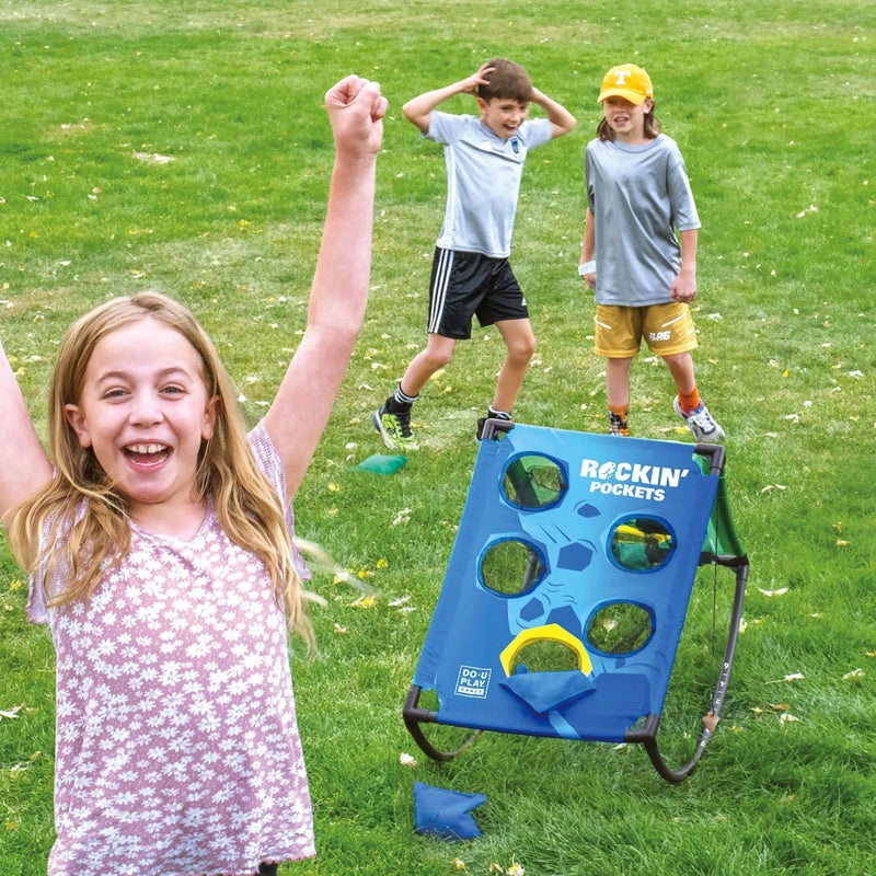 A girl in a pink floral shirt celebrates in the foreground with her arm raised, while two boys play on a blue Rockin' Pockets portable target game on a green grass lawn. The boy on the right throws a bean into the pockets.