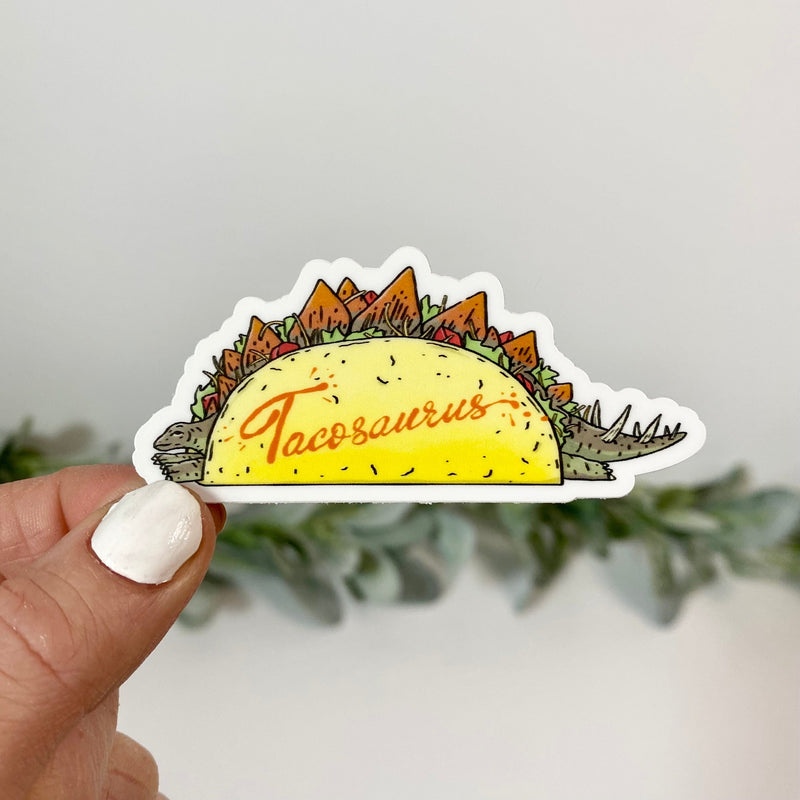 Hand holding Taco-saurus vinyl sticker showing stegosaurus shaped like taco with orange text