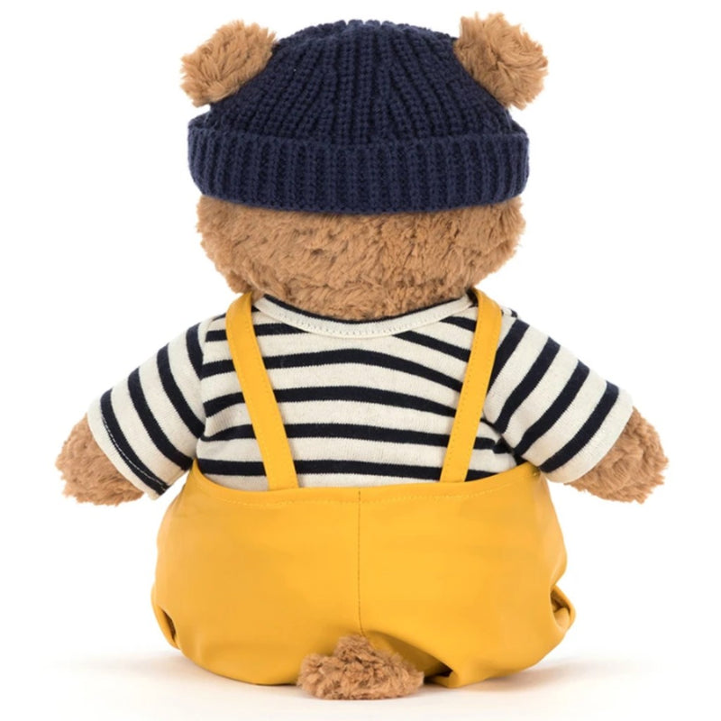Jellycat's Bartholomew Bear wearing a navy hat with pom-poms, striped shirt, and yellow overalls on a white background.