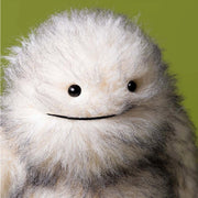A close-up of a fluffy white and gray plush toy with black eyes and a simple stitched smile, set against a solid olive green background.
