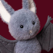 A closeup of the cute face of Jellycat's Broox Bat on a burgundy background.