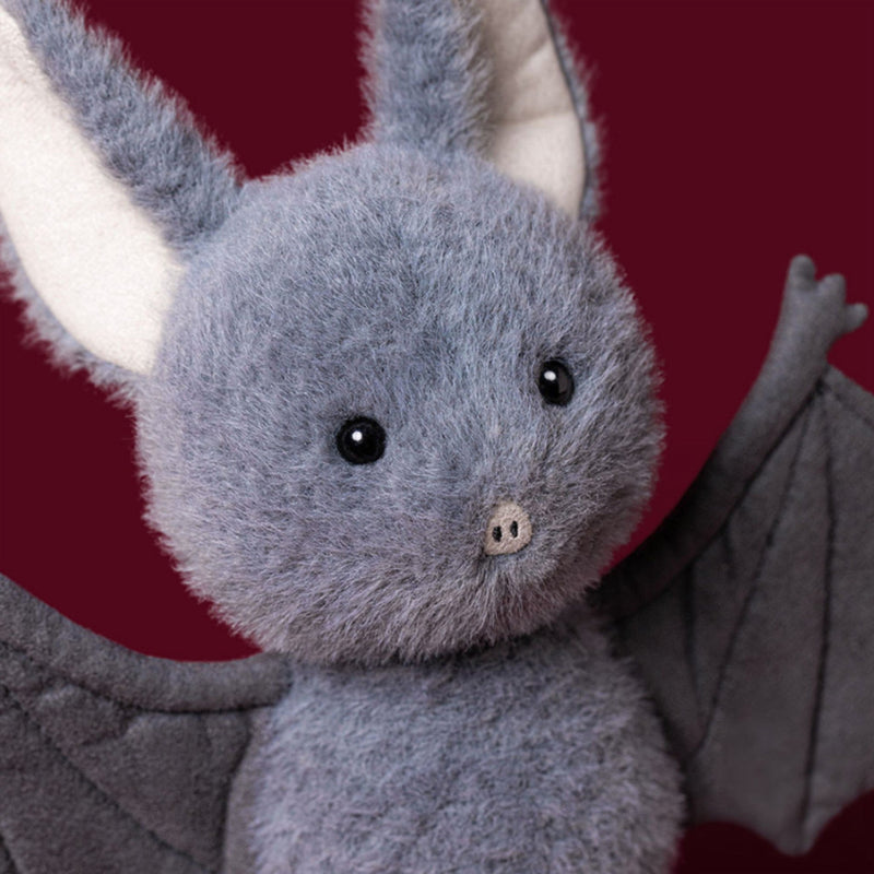 A closeup of the cute face of Jellycat's Broox Bat on a burgundy background.