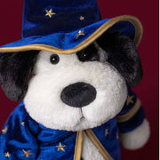 Jellycat's Rufferty dog plush dressed as a wizard, wearing a blue velvet hat and matching robe with gold star details, photographed against a dark red background.