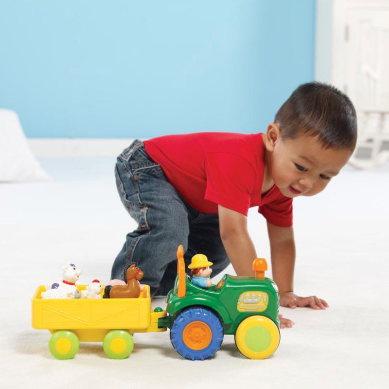 A young child in a red shirt and jeans crawls on a white floor while playing with a green and yellow toy tractor that pulls a yellow trailer filled with small animal figures.