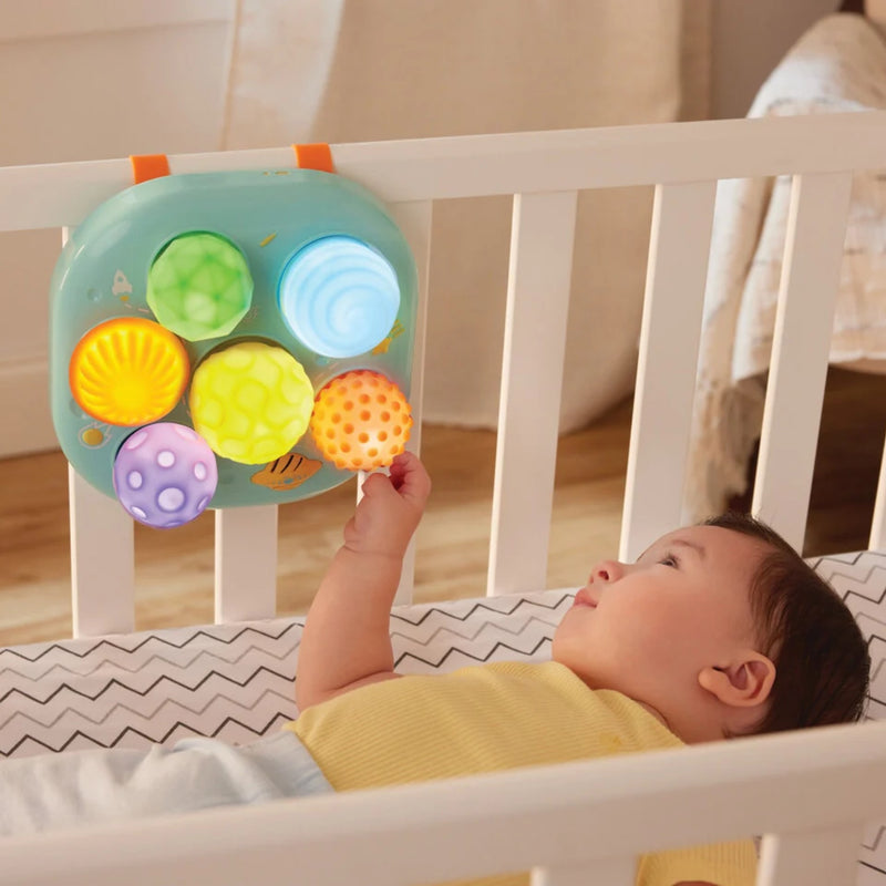 A baby lying in a white crib reaches toward a colorful baby activity toy hanging on the crib rail. The toy has a mint green base with orange handles and five textured, colorful balls in green, blue, yellow, orange, and purple.