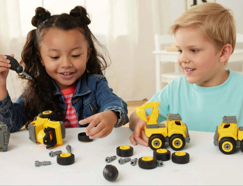 Two children playing with yellow construction toy vehicles and loose gray and black building pieces on a white table in a bright indoor room.
