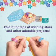 Hands folding a yellow paper star with tweezers, wearing colorful paper star rings. Above, an open box of folded paper stars and small folded paper creations are shown with the text 'Fold hundreds of wishing stars and other adorable projects!' on a light blue background with hanging paper stars.