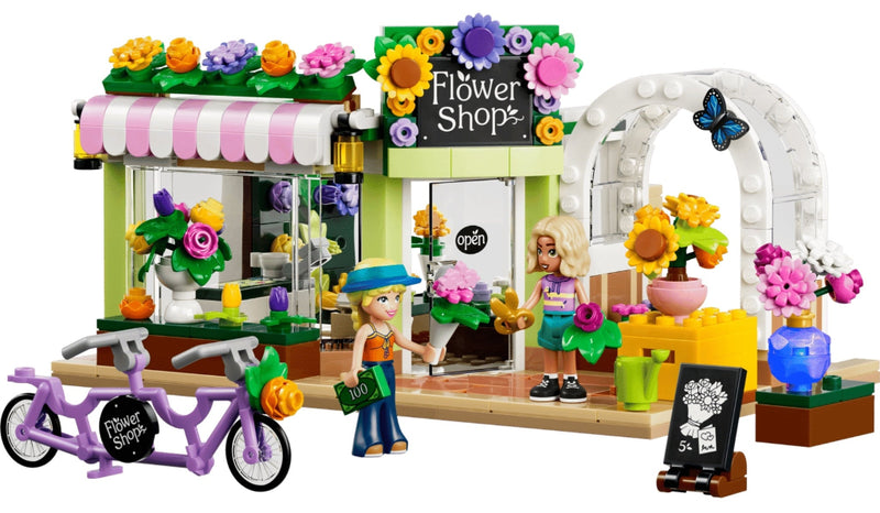 LEGO City Flower Shop set with a pastel green storefront, pink-and-white striped awning, flower boxes, a purple tandem bicycle, two minifigures, and a white arched entrance on a white background.