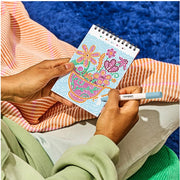 Person coloring a floral design in a small notebook with a pen.