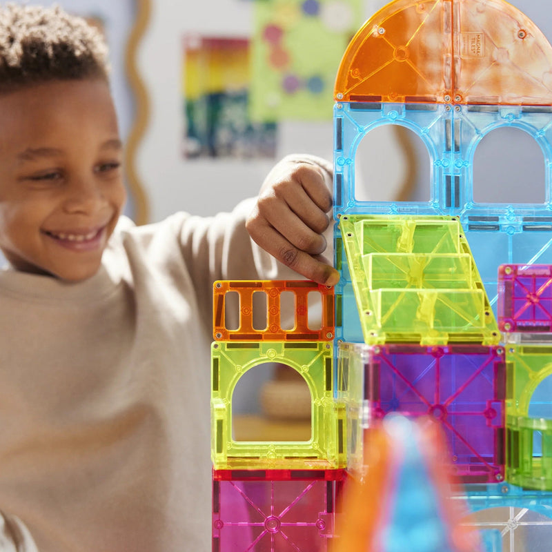 A young boy smiling and building a colorful translucent magnetic block tower with bright neon orange, blue, green, yellow, and purple pieces in a softly lit indoor setting.