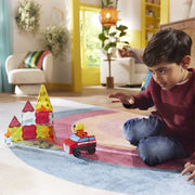 A child sits on a colorful rug in a bright living room, playing with a red toy car and a translucent magnetic block tower.