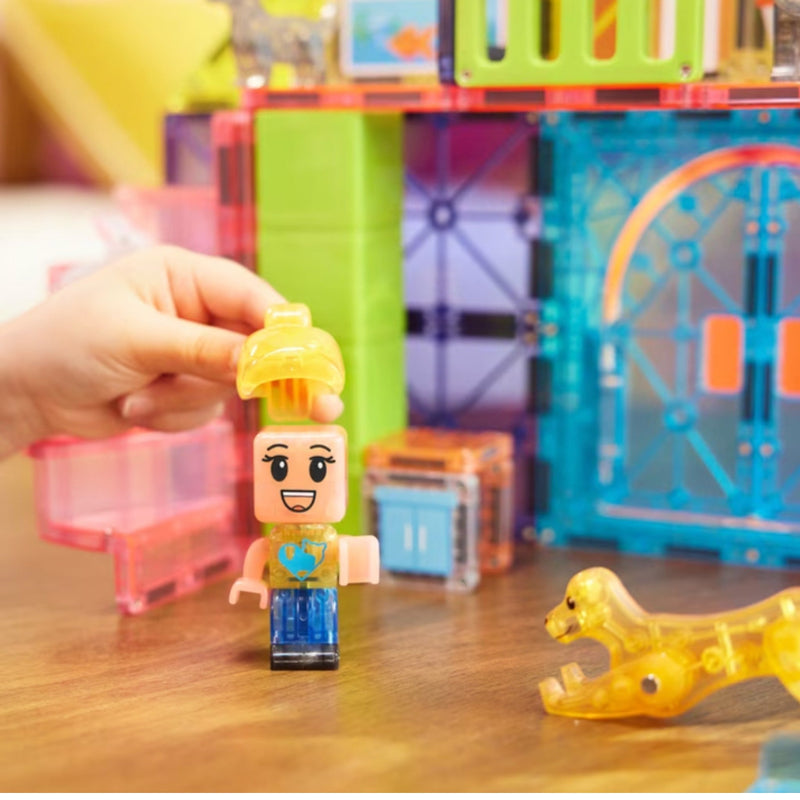 A child's hand is placing a small translucent building-block minifigure with a yellow construction helmet onto a colorful plastic toy building set on a wooden table. A translucent yellow dog figure is also visible nearby.
