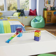Multicolor magnetic building blocks on a white table in a brightly lit children's room. The blocks include transparent acrylic pieces with embedded rainbow-colored rods and colorful solid tiles, arranged to form a small structure with a ramp.