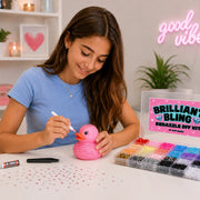 A girl in a blue t-shirt is sitting at a white table, beadzzling a pink rubber duck with a white marker. Next to her is a clear plastic case labeled 'Brilliant Bling Bedazzle DIY Kit' filled with colorful beads, and a pink glitter pen lies on the table.