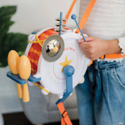 A child wearing a white top and blue jeans holds a colorful toy drum and cymbal set. The toy features a white drum face with a yellow star, orange patterned drum body, blue and orange straps, yellow maracas, and a blue mallet.
