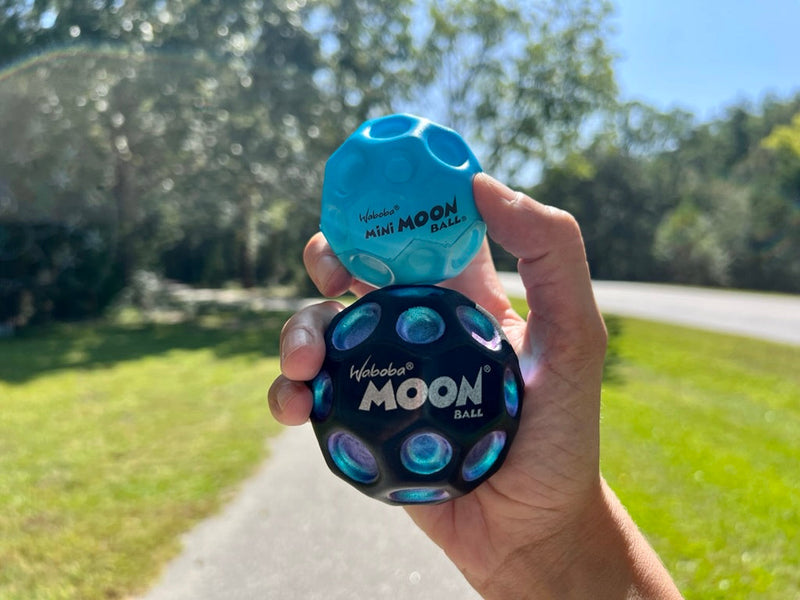 Hand holding two Waboba Moon Balls outdoors - one blue Mini Moon Ball and one black regular Moon Ball