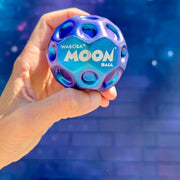 Hand holding Waboba Moon Ball in metallic blue and purple with crater-textured surface on blue background