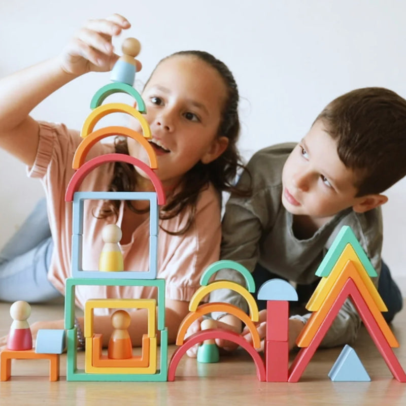 Two children playing with colorful wooden building blocks and small peg dolls on a light wood table against a plain light background.