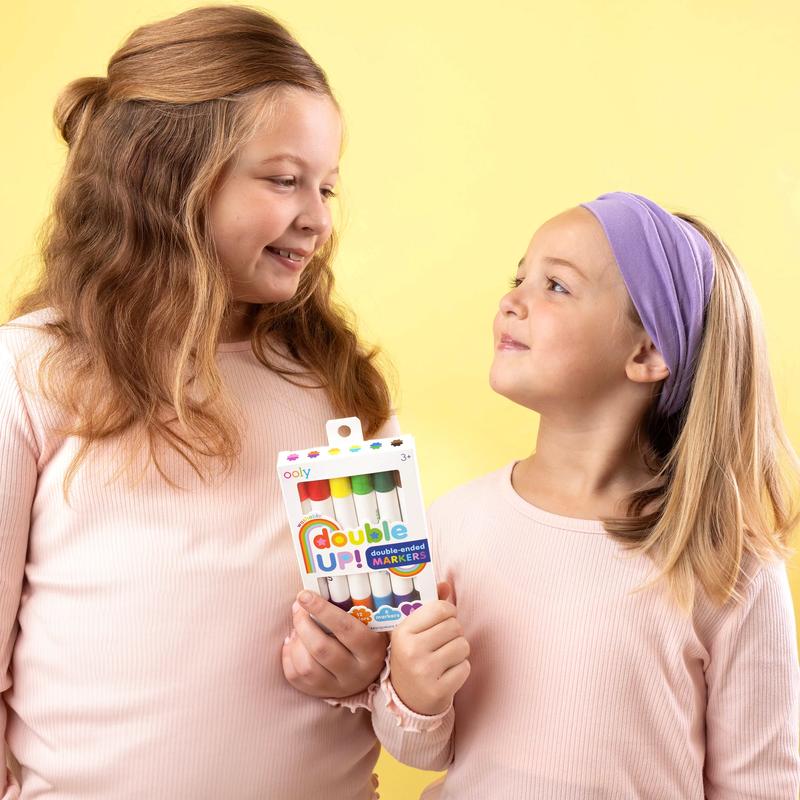 Two girls holding OOLY Double Up! Double Ended Markers package on yellow background
