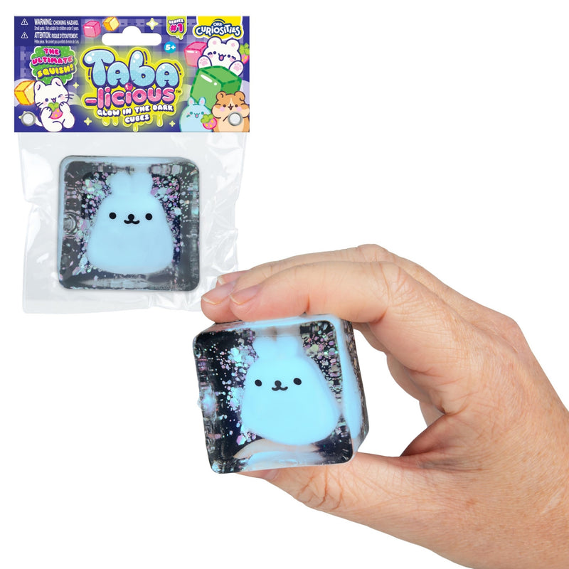 A hand holds a square squishy toy cube with a light blue fluffy bunny face and glittery black background. The package of the same product hangs above on a plain white background.