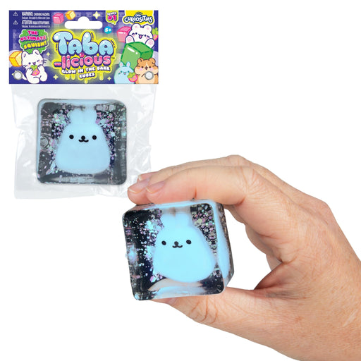 A hand holds a square squishy toy cube with a light blue fluffy bunny face and glittery black background. The package of the same product hangs above on a plain white background.
