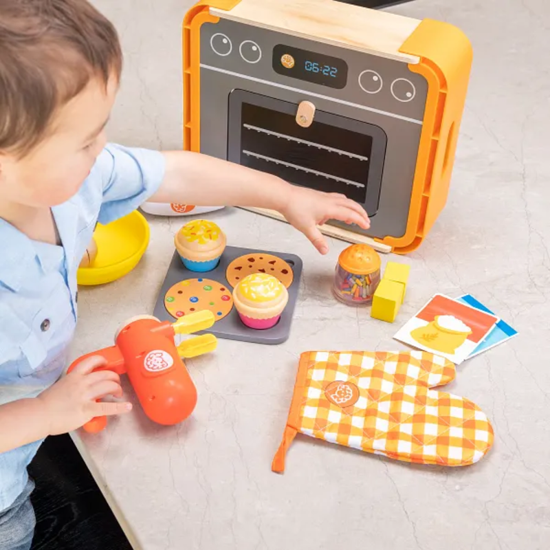 Child playing with a toy baking set including an oven, muffin tray, and utensils on a light surface.