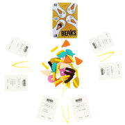 The Beaks card game set arranged on a white background, featuring a yellow tin with the Beaks logo, several cream-colored play money cards, and a central pile of colorful food-shaped tokens.