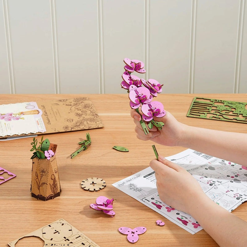 Person crafting a pink flower arrangement on a wooden table with craft materials.