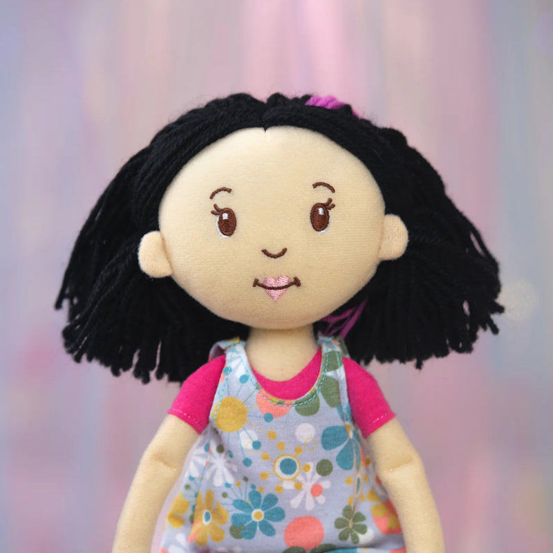 Close up of the Daisy Groovy Girls plush doll with black hair and a colorful dress against a soft pink background.