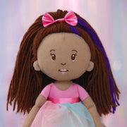 Close-up of Groovy Girl Iris plush doll with brown hair, pink bow, and pink and blue dress on a soft pink background.