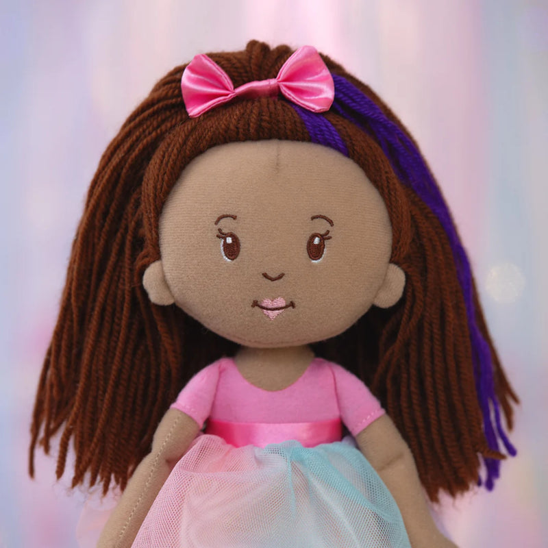 Close-up of Groovy Girl Iris plush doll with brown hair, pink bow, and pink and blue dress on a soft pink background.
