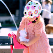 Child wearing a colorful helmet and a teddy bear into her Scootaseatz, standing next to a pink scooter.
