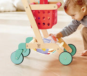 Child playing with wooden Hape shopping cart toy with red basket and green wheels