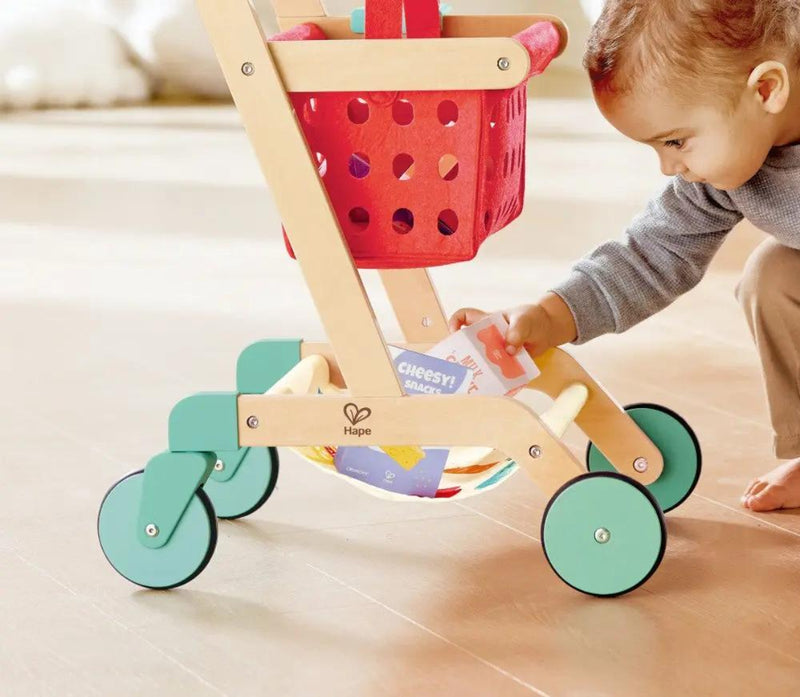 Child playing with wooden Hape shopping cart toy with red basket and green wheels