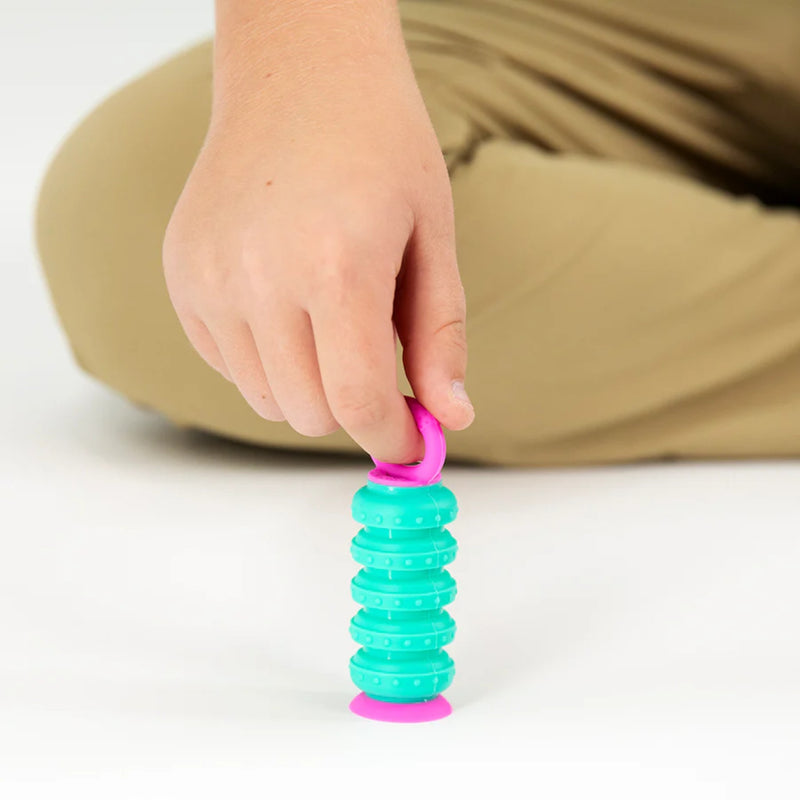 A close-up of a person's hand holding a bright pink Krumple with a ring handle above a teal and pink coiled toy on a white surface, with tan pants blurred in the background.